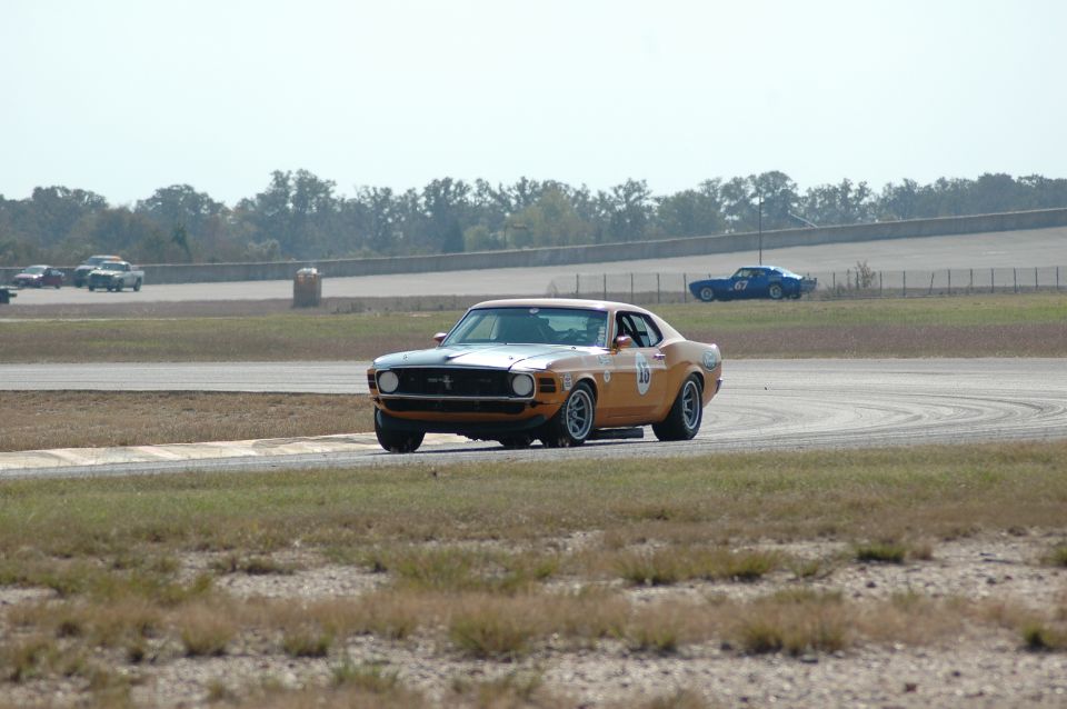 Orange Mustang race car turning in at a corner with a blue No. 67 car in the distance