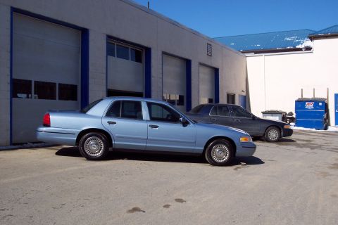 2001 Ford Crown Victoria - Two sedans, one light blue and one dark, are parked in front of a commercial building with large garage doors.