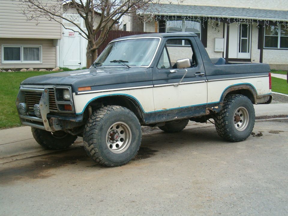 Lifted black and white 1980s Ford Bronco with top removed behind cab, on big mud tires, parked on street