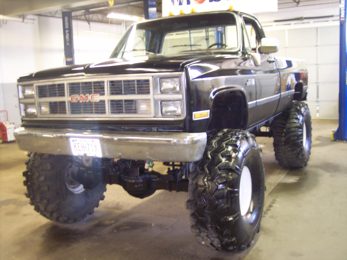 Black lifted square-body GMC pickup on tall mud tires and white wheels inside a shop