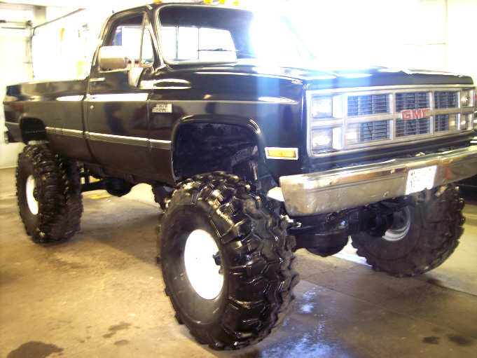 Black lifted GMC pickup with large mud tires and white wheels, parked indoors.