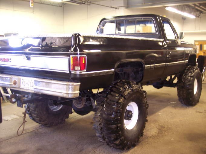 Black lifted GMC pickup, rear 3/4 view, on large mud tires indoors