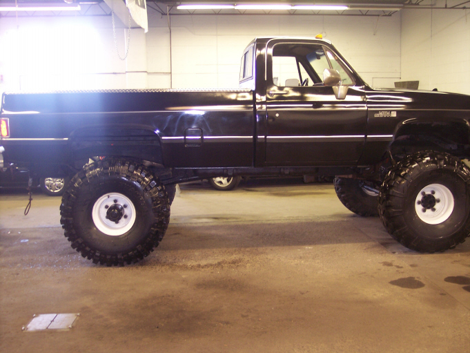 Black lifted single-cab pickup on large mud tires, side view in a garage.