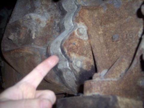 1984 GMC Sierra 3500 - A finger points to a seam on a heavily rusted metal component, likely part of a vehicle's drivetrain.