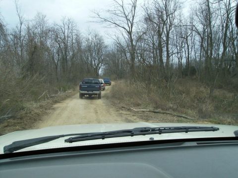 1988 Ford Bronco - Three trucks travel down a dirt road surrounded by bare trees.
