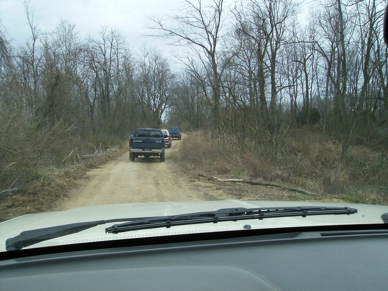 View from inside a vehicle looking at three trucks driving on a dirt road through a wooded area