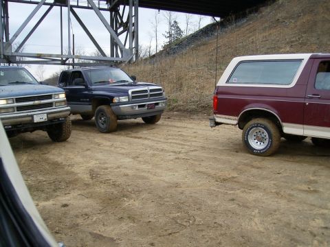 1988 Ford Bronco - Three trucks are parked on a dirt road under a large bridge structure.