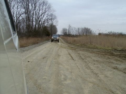 1988 Ford Bronco - A gray SUV drives down a dusty rural road, with dry grass and trees lining the path under a cloudy sky.