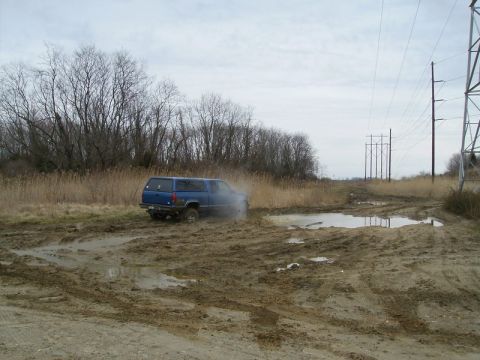 1988 Ford Bronco - A blue SUV with a camper shell navigates a muddy track, with tall grass and power lines in the background.