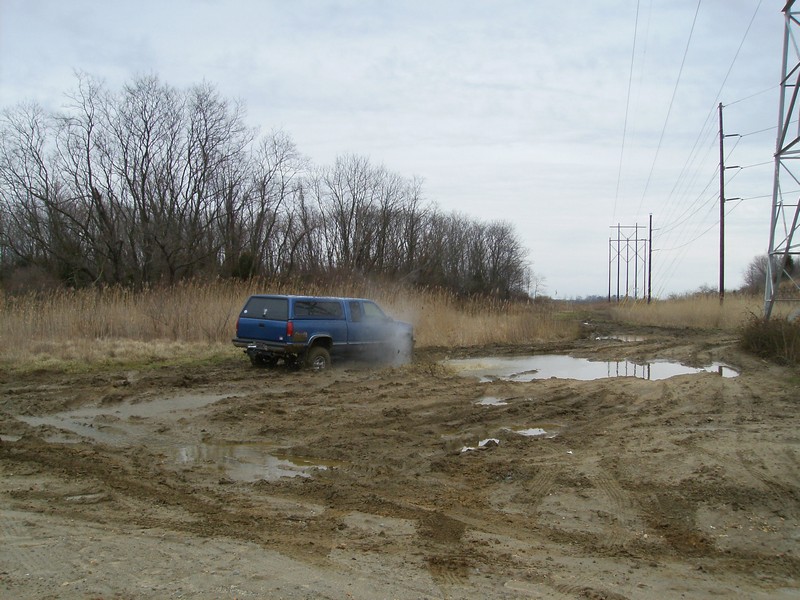 Blue SUV with camper shell driving through mud, kicking up spray