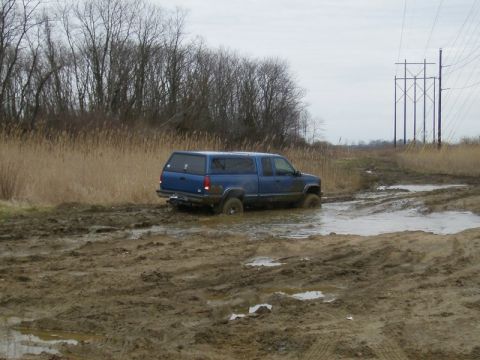 1988 Ford Bronco - Blue pickup truck with a camper shell navigates a muddy off-road trail.
