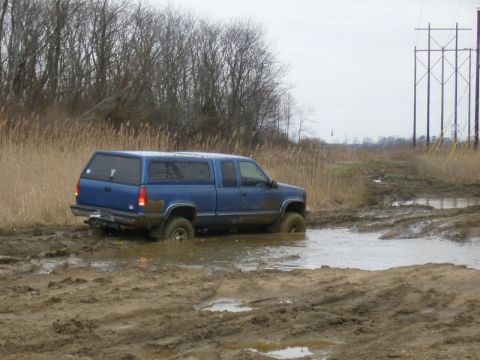 1988 Ford Bronco - A blue pickup truck navigates a muddy terrain, its tires submerged in water and mud.