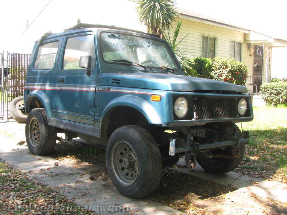 Teal 1987 Suzuki Samurai parked in a driveway, front grille missing, windshield cracked