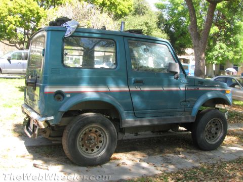 1987 Suzuki Samurai - A teal Suzuki Samurai is parked on a paved surface with trees in the background.