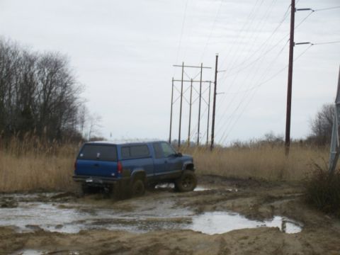 1988 Ford Bronco - A blue pickup truck with a camper shell navigates a muddy track under an overcast sky.