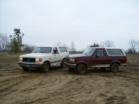 1988 Ford Bronco - Two Ford Broncos sit on a muddy trail under an overcast sky, with bare trees dotting the landscape.