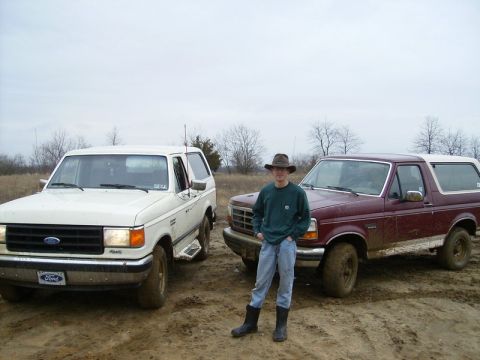 1988 Ford Bronco - Two Ford Broncos are parked in a muddy outdoor area with a person standing between them.