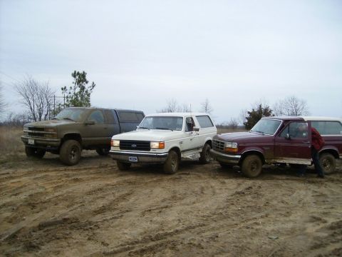 1988 Ford Bronco - Three trucks and SUVs are parked in a muddy outdoor area under an overcast sky.