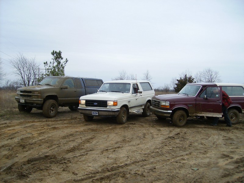 Three trucks and SUVs parked in a muddy area. One is a white Ford Bronco, another is a maroon Ford F-series truck, and the third is a dark green Chevrolet pickup