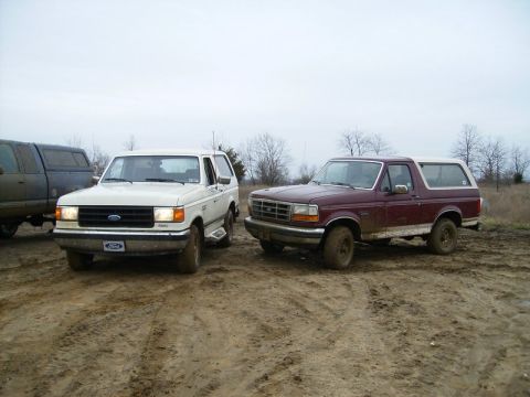 1988 Ford Bronco - Two Ford Broncos, a white one and a maroon one, are parked side-by-side on a muddy surface.