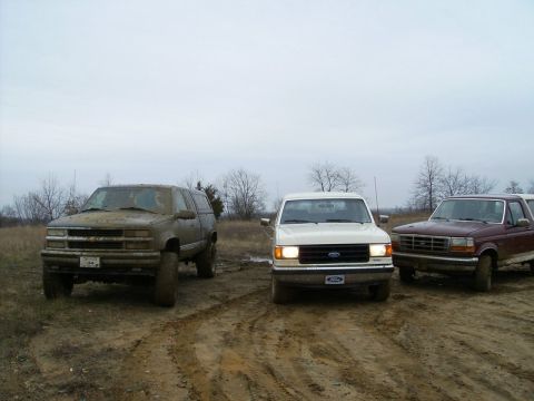 1988 Ford Bronco - Three trucks, including a maroon Ford Bronco and a white Ford F-Series, are parked in a muddy off-road area.
