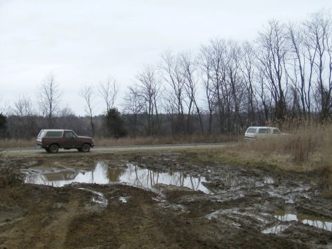 1988 Ford Bronco - Two SUVs navigate a muddy track with puddles and bare trees in the background.