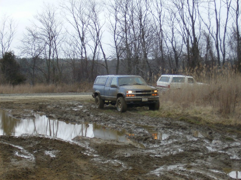 Blue Chevy pickup truck with a cap driving through a muddy field. Another white SUV is visible in the background