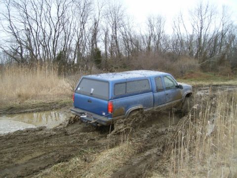 1988 Ford Bronco - A blue pickup truck with a camper shell navigates a muddy off-road course, getting stuck in the mire.