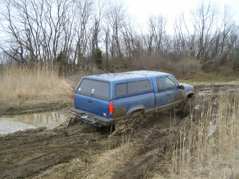 Blue pickup truck with camper shell stuck in mud, rear 3/4 view