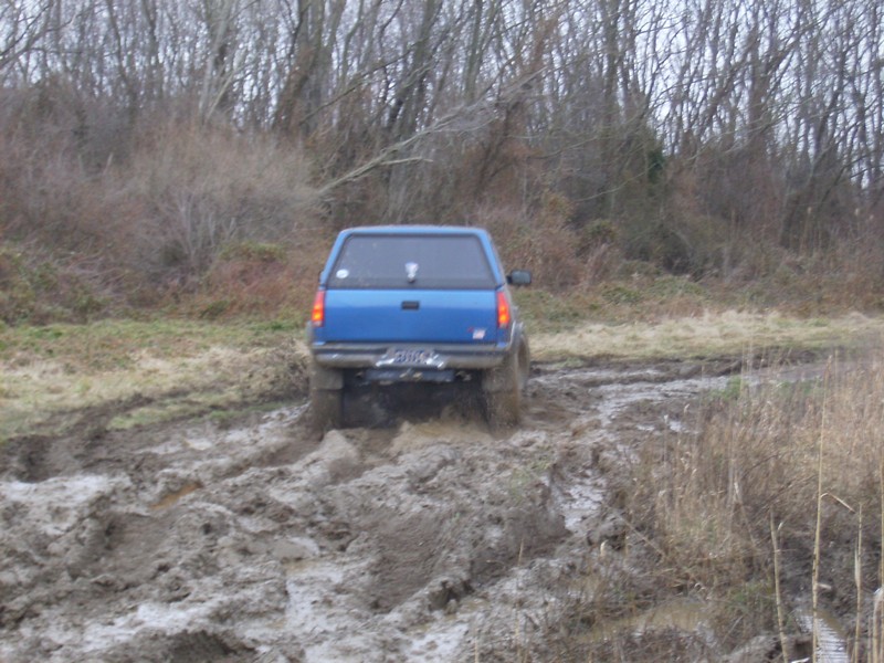 Blue pickup truck with a cap driving through a muddy trail with trees in the background