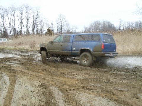 1988 Ford Bronco - A mud-splattered blue GM extended-cab pickup with a matching bed cap slings water and mud from its rear tire while running through a rutted…