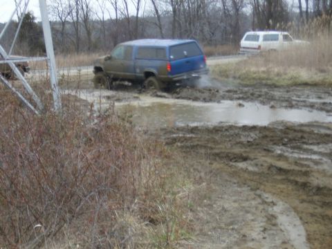 1988 Ford Bronco - A blue extended-cab pickup with a matching bed cap plows through a wide mud pit, throwing spray behind its rear tires, with mud caked over…