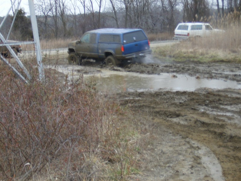 Blue pickup with bed cap running through a wide muddy field, spray behind tires