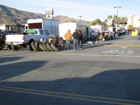 1986 Ford Bronco - A group of people stand around a pickup truck and tires at an outdoor event, with trailers and mountains in the background.