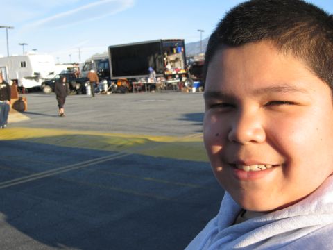 1986 Ford Bronco - A boy smiles for a selfie at a swap meet with various vehicles and vendor stalls visible.