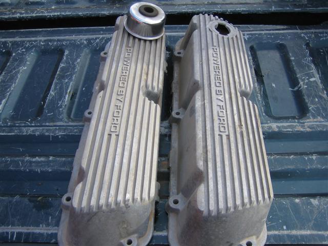 Two aluminum valve covers with "POWERED BY FORD" cast into them, resting on a dark textured surface