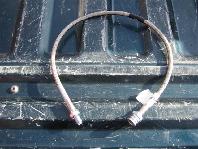 Stainless steel braided brake hose with black heat shrink and metal fittings, lying on a dark green textured surface