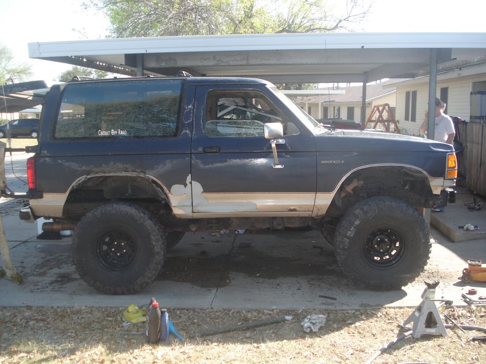 Side view of a dark blue Ford Bronco II with large off-road tires and some body damage. It has a tan stripe and a sticker on the rear window