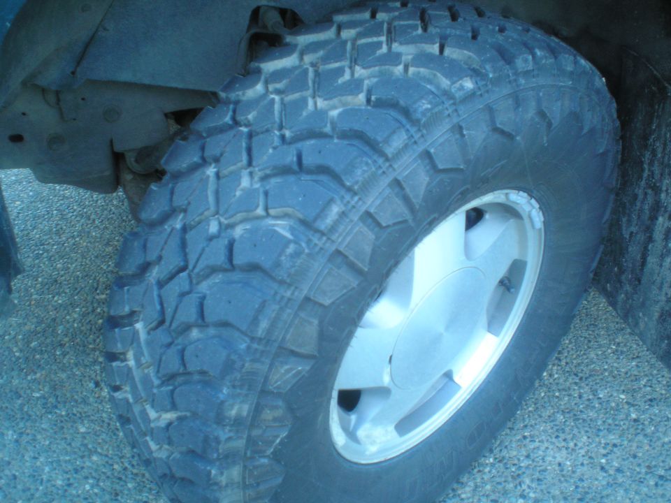 Close-up of a knobby off-road tire on a silver alloy wheel, with visible suspension components above