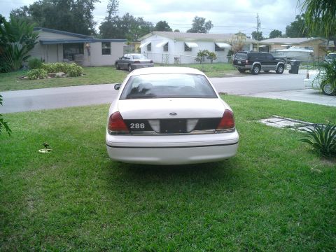 1999 Ford Crown Victoria - A white sedan is parked on a grassy lawn in front of a residential house, with other vehicles in the background.