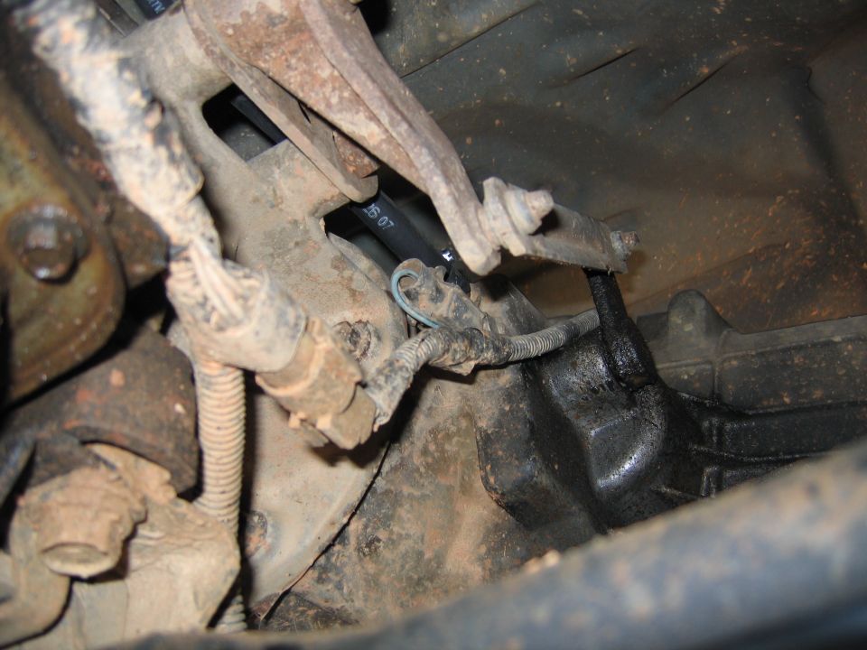 Close-up under a vehicle showing rusty parts, wiring harness, spring-wrapped line, and oily black component