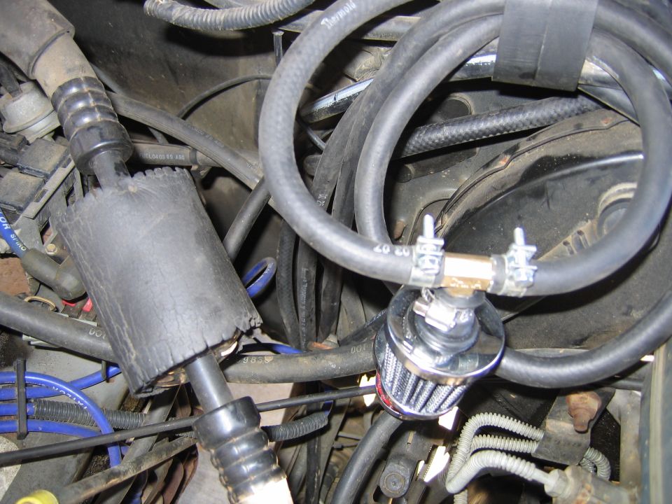 Engine bay close-up with small chrome breather filter clamped to a black hose among wires and hoses