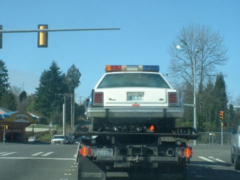 1987 Ford Crown Victoria LTD - Police car being towed on a flatbed truck, seen from behind at an intersection.