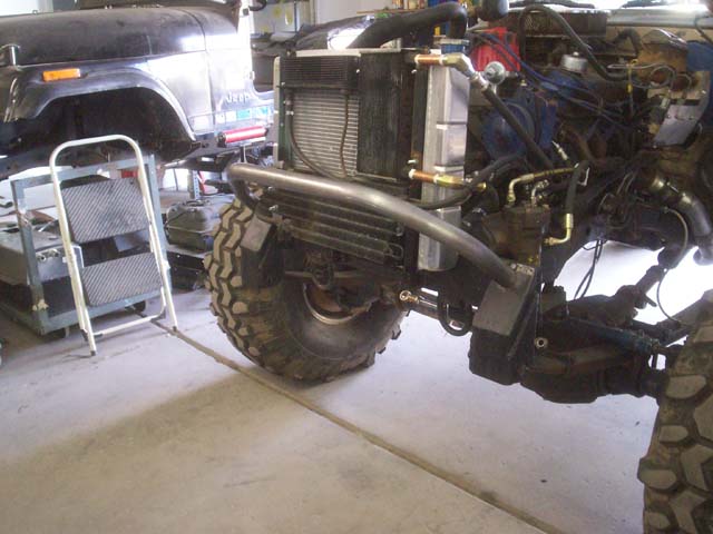 Off-road vehicle engine bay with radiator, hoses, and suspension components visible. Another vehicle is partially seen in the background