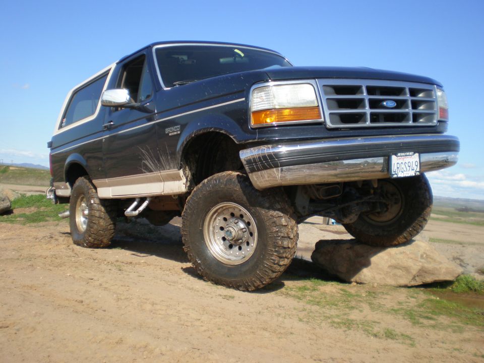 Dark blue 1993 Ford Bronco with a white top, lifted on large tires, driving over rocks