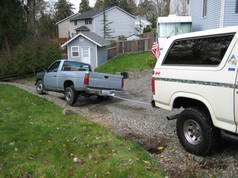 1980 Ford Bronco - A Nissan pickup truck is being towed by a Ford Bronco, with houses and trees in the background.