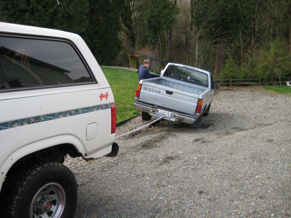 White Ford Bronco towing a silver Nissan pickup truck on a gravel driveway