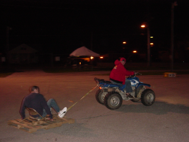 At night, ATV tows a person sitting on a wood pallet sled across a parking lot