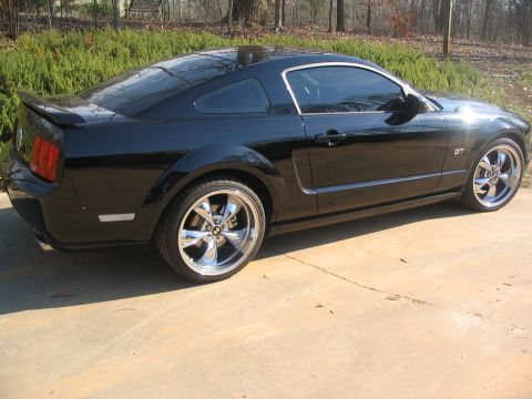 2007 Ford Mustang - A black Ford Mustang coupe is parked on a concrete surface with a wooded background.
