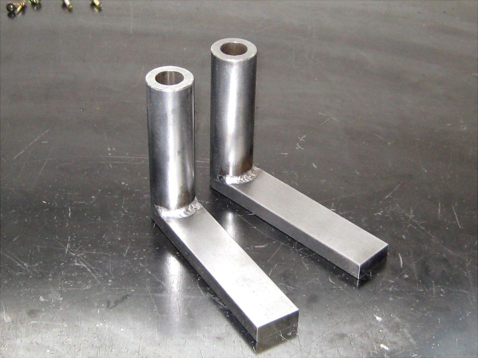 Two L-shaped metal brackets with cylindrical extensions, welded together, resting on a scratched metal surface